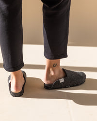 On a light floor, someone in dark pants wears gray slipper shoes by graf lantz. One slipper, crafted from sustainable Merino wool felt, is partially removed, revealing a horseshoe tattoo on their right ankle. Soft sunlight casts shadows across the floor.