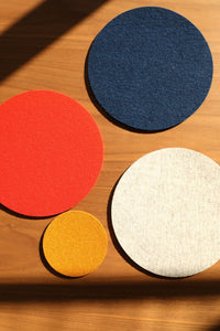 Four circular graf lantz coasters made of Merino wool in blue, red, beige, and yellow rest on wood. Sunlight and shadow create diagonal lines across them. These Oeko-Tex® Certified pieces add style and function to your table setting.