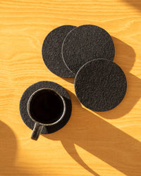 A black coffee cup is on a Bierfilzl Merino Wool Round Coaster with three more black coasters skillfully placed nearby on a light wooden table. Sunlight casts gentle shadows, accentuating the beautiful texture of these fine German Merino wool coasters.
