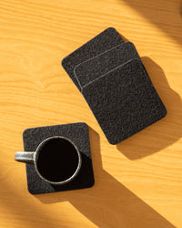A small black cup of coffee rests on a square Beer Felt coaster from graf lantz atop a wooden surface, with three more coasters stacked nearby casting shadows in the warm sunlight.