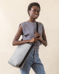 A person smiles in a sleeveless gray top and light blue jeans, holding a graf lantz Merino Wool Tote. The totes everyday functionality is matched by its sustainable design, featuring a gray color with a black strap against a soft beige background.