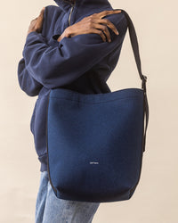 A person in a navy sweatshirt holds a large Merino Wool Tote by graf lantz, praised for everyday functionality. Its simple design with subtle text pairs perfectly with light blue jeans, all against a minimal, neutral-toned background.