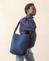 A person smiles while holding a blue tote bag from graf lantz over their shoulder, showcasing everyday functionality. Theyre wearing a navy merino wool sweater and jeans against a plain beige background.