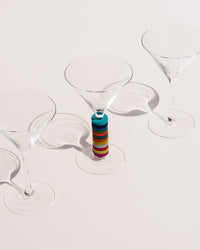 Three martini glasses sit on a light surface. The middle one is adorned with colorful Wine-Os from graf lantz, made of Bavarian Merino wool felt, around its stem, casting long shadows.