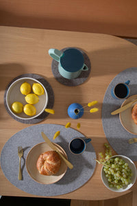 A table setting features croissants, yellow lemons in a bowl, grapes, and two cups of coffee. In the center is a blue and green jug, with a small blue vase of yellow flowers nearby. Everything rests on a wooden table enhanced by elegant graf lantz Merino Wool Placemats.
