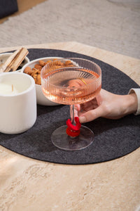 A hand holds a glass of rosé wine with a red glass marker from graf lantz on the stem, resting on a round grey placemat. Nearby, a white candle and a bowl of almonds create an eco-friendly luxury scene on a light wooden table.