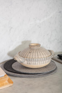A ceramic pot with a ribbed lid sits atop graf lantzs moisture-wicking Merino wool felt pads on a marble countertop. A light marble wall serves as the backdrop, accompanied by part of a wooden chopping board.