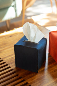 A navy blue graf lantz tissue box, with a minimalist silhouette, rests on a wooden table. A white tissue is partially pulled out, while sunlight highlights the tables texture. A hint of a red object is visible nearby.