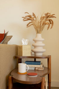 A cozy side table with two tiers features a wavy beige vase of dried grasses, books, a tan tissue box, and a white mug resting on a graf lantz Merino wool coaster. The scene beside the light couch exudes warmth and invites relaxation.