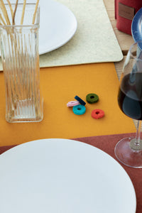 A table setting showcases white plates and red wine on a mustard-colored graf lantz placemat, featuring colorful felt rings as glass markers. Tall glass vases with breadsticks add to the sustainable luxury of this textured and vibrant decor.