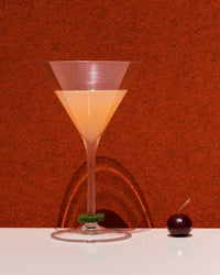 A tall, triangular graf lantz cocktail glass containing a peach-hued drink sits on a white surface against a bold red backdrop. wine glass markers and a single cherry nearby create clear shadows.