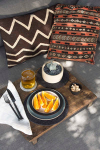 Orange slices and almonds on a wooden table accompany a glass of beer and black cutlery on a graf lantz heat-resistant napkin. A small plant adds greenery, while two cushions with zigzag and geometric patterns cast soft shadows over the cozy setting.