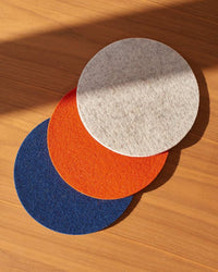 Three Merino wool felt pads from graf lantz, in gray, orange, and blue, are arranged on wood with a slight overlap. Sunlight casts a diagonal shadow across them, accentuating the texture of the moisture-wicking felt.