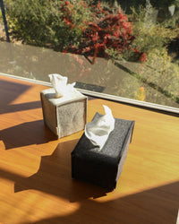 Two graf lantz tissue boxes with sustainable fabric covers, one light gray and one dark gray, rest on a wooden table by the window. The sun casts shadows on the table, while outside, red-flowered bushes and greenery are visible.