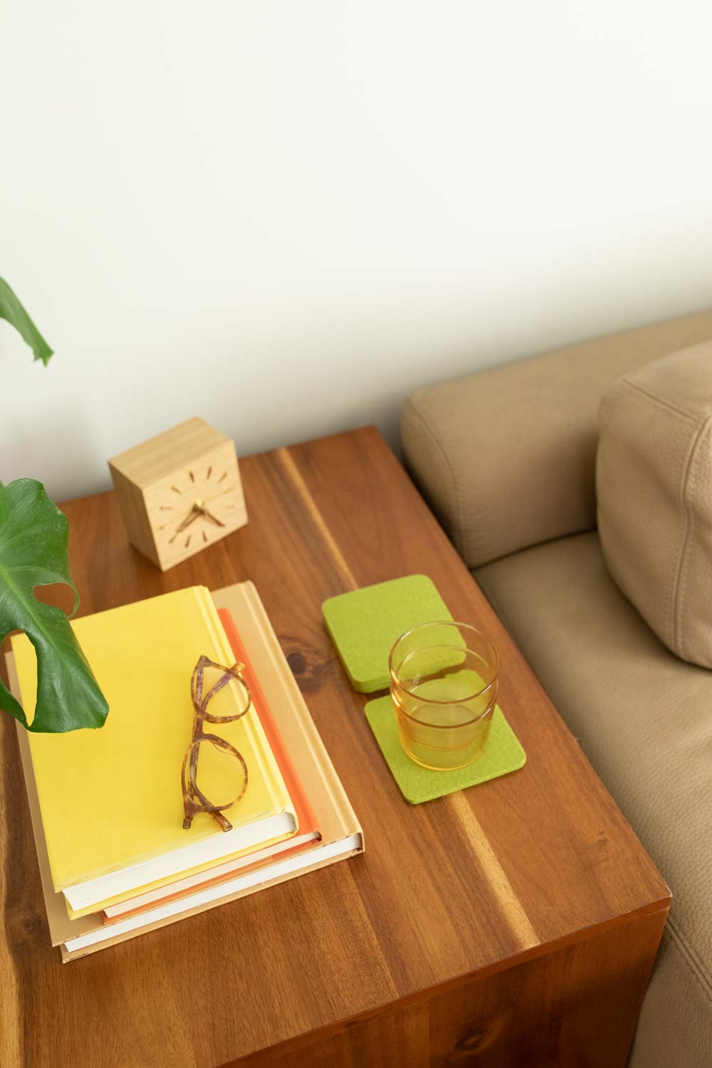 A sleek wooden side table by graf lantz, adorned with stacked books, glasses, a small wooden clock, and an elegant green coaster under a glass of yellow liquid, enhances the beige couch and large green leaf backdrop for seamless home décor sophistication.