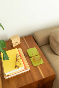 A sleek wooden side table by graf lantz, adorned with stacked books, glasses, a small wooden clock, and an elegant green coaster under a glass of yellow liquid, enhances the beige couch and large green leaf backdrop for seamless home décor sophistication.