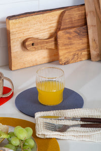A glass of orange juice sits on a gray heat-resistant coaster by graf lantz, placed on a white surface. Wooden cutting boards and a plate with grapes and bread are nearby, along with a fork and knife on a white napkin.
