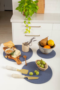 A modern kitchen counter showcases a graf lantz cutting board with nuts, cheese, and cheese knives on round blue placemats, a small vase of lavender, a bowl of pears and oranges, and green grapes on a dish. A plant hangs above white cabinets in this heat-resistant display of lasting elegance.