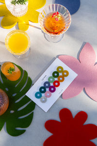 A table features graf lantz’s eco-friendly, felt leaf-shaped placemats and drinks alongside a white card with colorful rings, possibly for Wine-Os markers. The sustainable wool design casts charming shadows, enhancing the elegance.