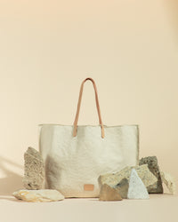The graf lantz beige canvas tote, with its premium vegetable-tanned leather handles, is an upscale market bag. Set against a warm, light tan background with various rocks, it embodies earthy elegance and enhances its neutral tones and luxurious appeal.