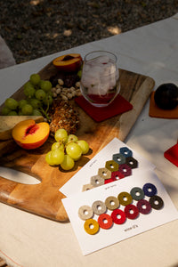 A graf lantz wooden cutting board elegantly displays grapes, peaches, nuts, and a mixed seeds roll. A wine glass with ice sits on a red coaster with wine glass markers nearby. Two color sample cards labeled grit luts and wine os are placed on the tablecloth.