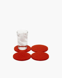 A glass of water with ice rests on an orange Merino wool coaster by graf lantz. Three more coasters surround it closely on a white background, enhancing the scenes warmth and elegance.