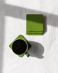 A green ceramic cup with dark liquid rests on a graf lantz Beer Felt coaster on a white textured surface. Beside it is a stack of identical coasters. Sunlight casts soft shadows across the scene.