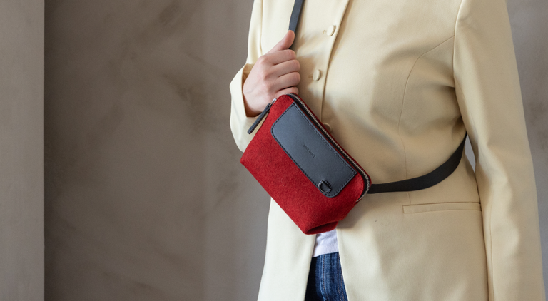 A person in a light beige outfit holding a brown crossbody bag with a beige flap, adjusting the strap. The surroundings are a neutral, soft background.