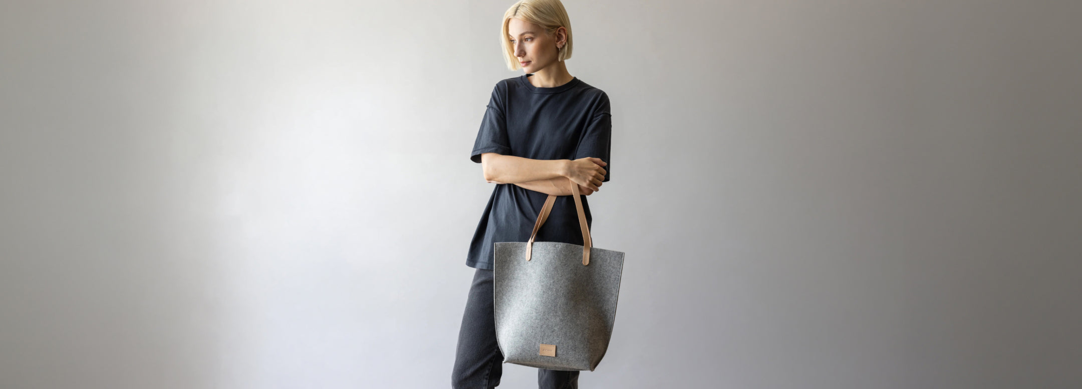 Person in a long-sleeved, cream-colored dress holding a gray, perforated handbag with a wooden handle, standing against a textured rock background.