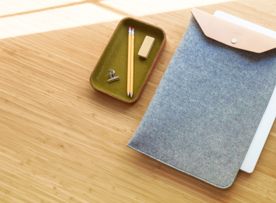 A felt sleeve with a leather flap lies next to a tray holding two pencils, an eraser, and a metal binder clip on a light wooden surface in sunlight.
