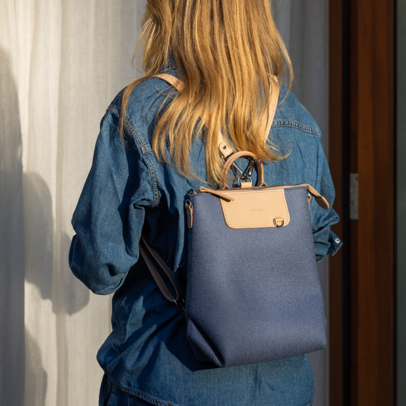 A person with long blonde hair wears a blue denim shirt and a blue backpack with a tan flap, standing in sunlight near a curtain and a wooden door.
