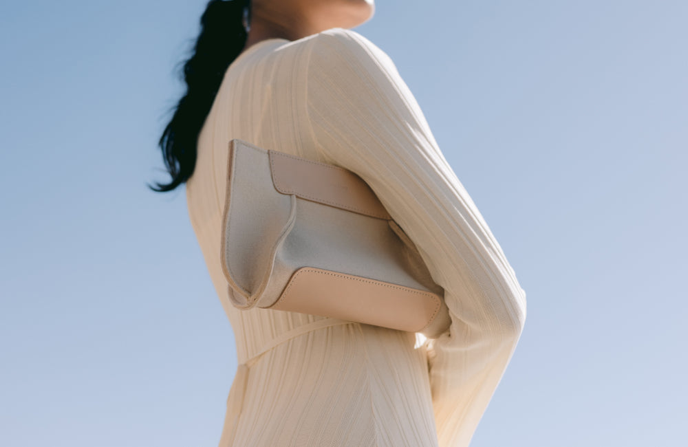 A woman in a light, long-sleeved dress holds a beige clutch purse against her side, standing against a clear blue sky. Her face is out of frame, and her dark hair is tied back.
