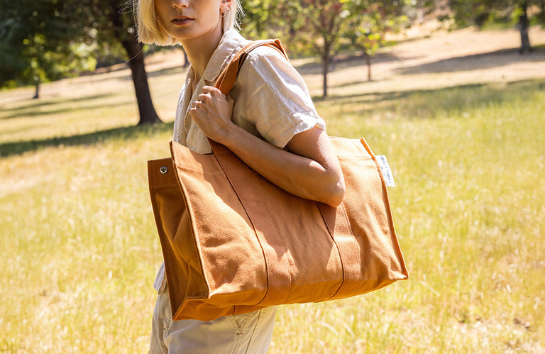 A person with short blonde hair carries a large brown canvas tote bag over their shoulder while standing in a sunlit grassy field with trees in the background.