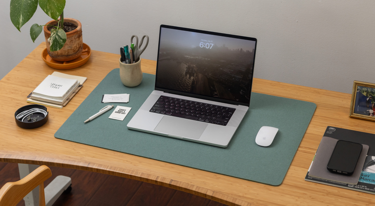 Person in a white shirt typing on a laptop at a wooden desk. A smartphone and a glass of water are placed nearby. A window with leafy greenery is in the background.