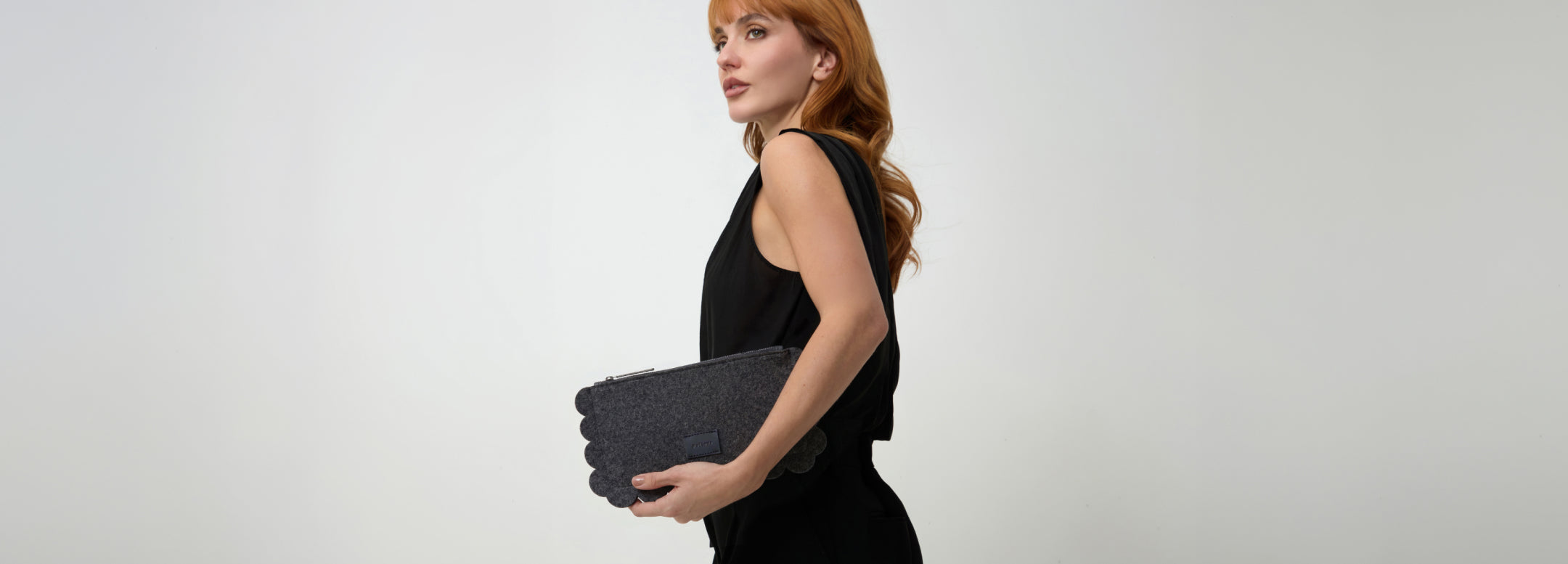 A woman in an off-the-shoulder cream dress sits on a rock against a light wall, placing a cellphone into a tan handbag. A leafy branch casts a shadow nearby.