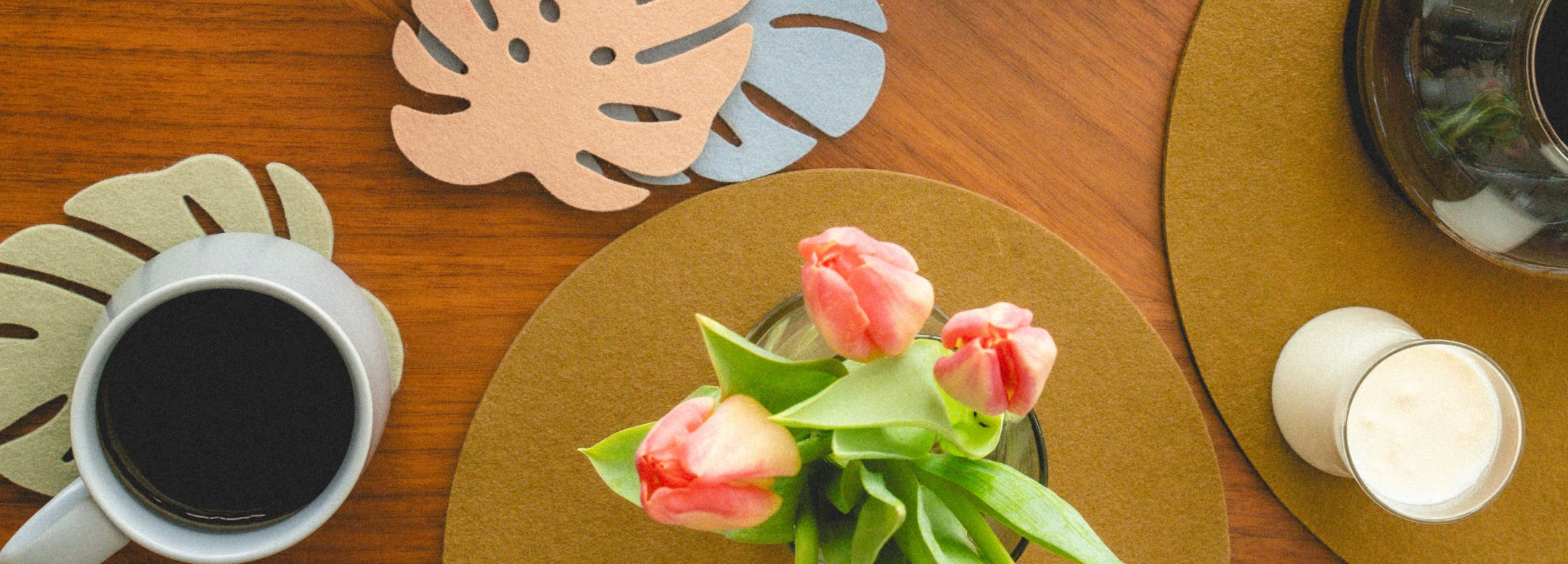 A cup of black coffee, a vase with pink tulips, a candle, and leaf-shaped coasters are arranged on a wooden table with a round brown placemat.