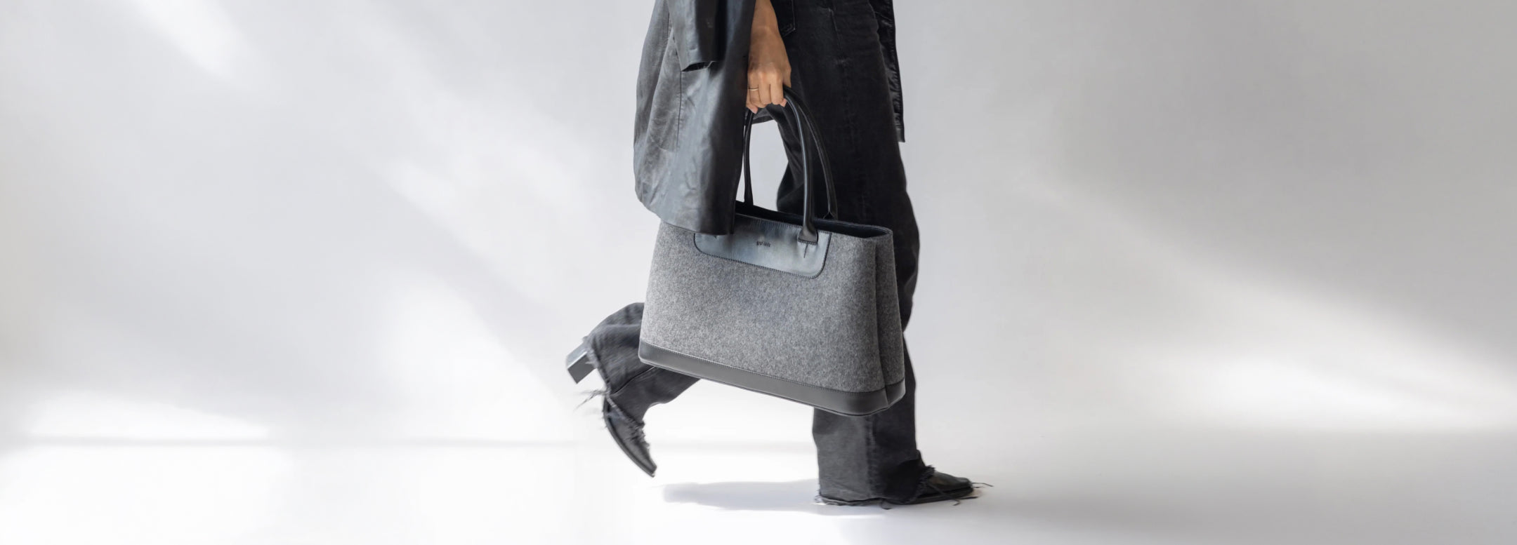 A person dressed in dark clothing walks while holding a large gray tote bag with black handles against a plain, light background.