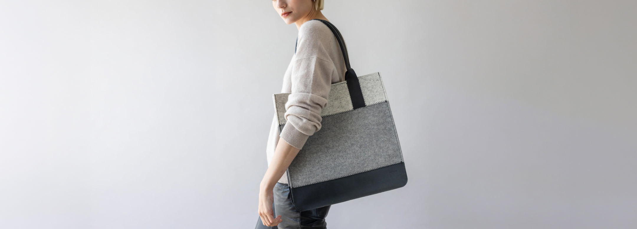 A woman in a dark green suit and glasses stands in profile, carrying a large, stylish two-toned tote bag with black handles against a light textured wall.