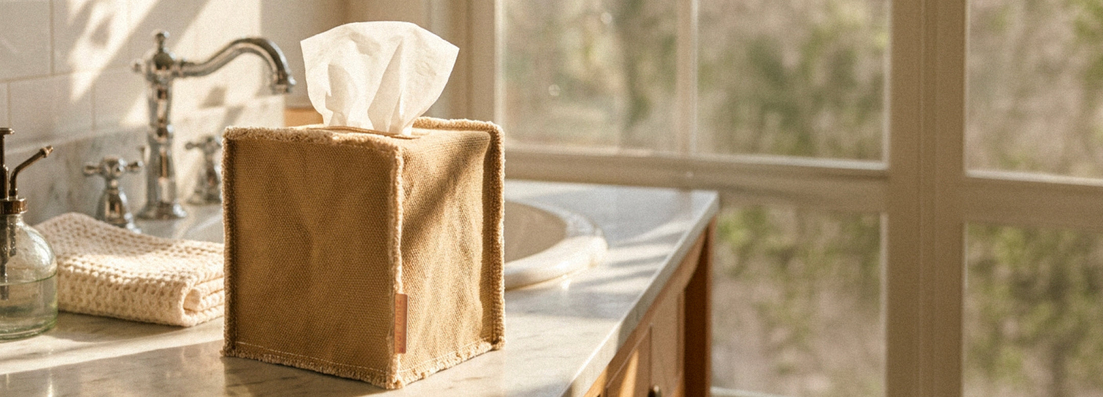 A grey felt tissue box sits on top of a book titled MARK BRADFORD, placed on a wooden table with blurred objects in the background.