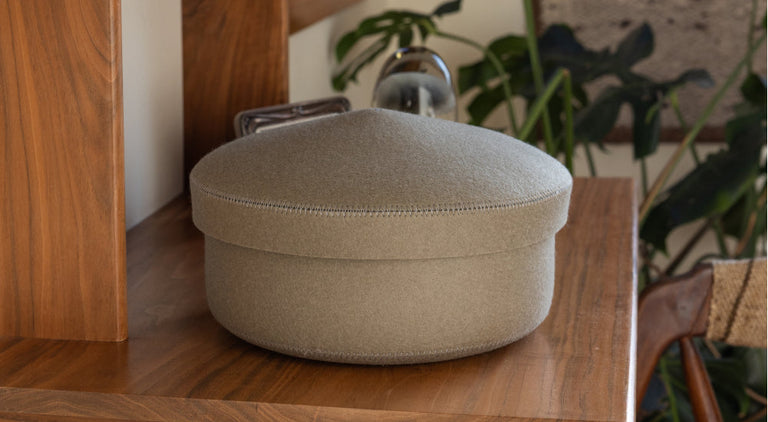 A round, light gray fabric storage box with a lid sits on a wooden shelf, surrounded by leafy green plants and natural decor elements.