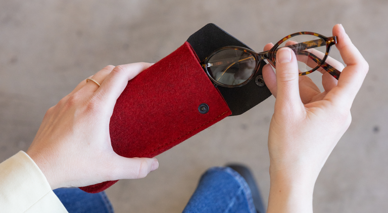 A person in a white, long-sleeved top holds a black pouch in one hand and a small bottle of peach-colored makeup in the other, standing against a rocky background.