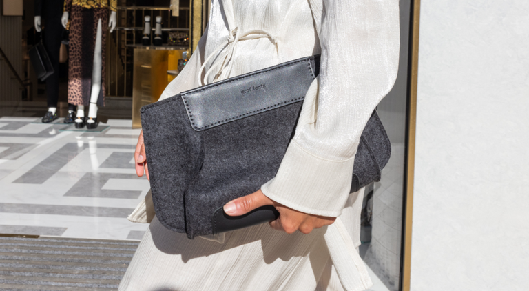 A woman in an off-the-shoulder cream dress sits on a rock against a light wall, placing a cellphone into a tan handbag. A leafy branch casts a shadow nearby.