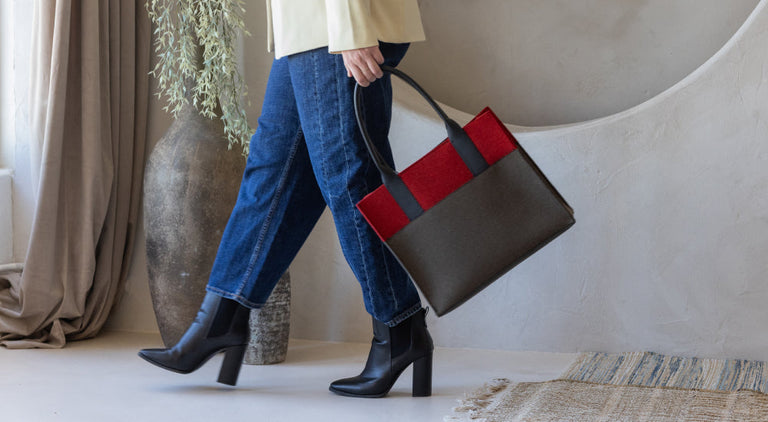 A person in blue jeans, black heeled boots, and a light blazer carries a stylish brown and red handbag indoors near a large vase and a woven rug.