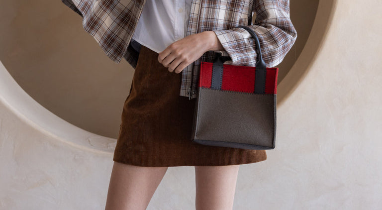 A person wearing a plaid shirt, white blouse, and brown skirt holds a stylish gray handbag with red and black accents against a beige background.
