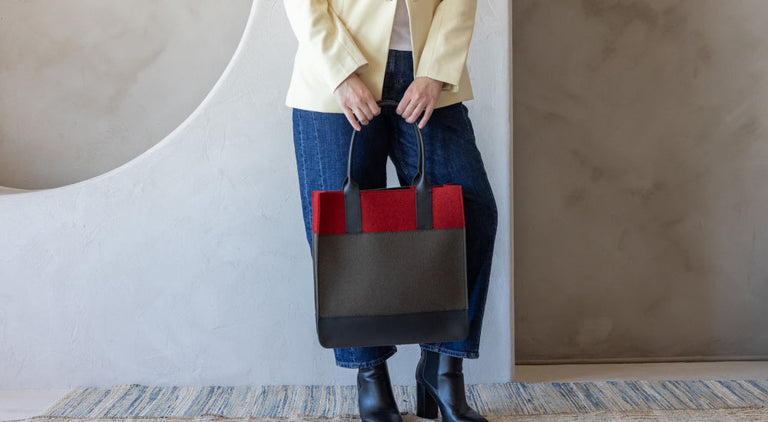 Person in blue jeans, black boots, and a cream blazer holding a large color-block tote bag with red, dark gray, and black panels, standing on a woven rug against a textured wall.