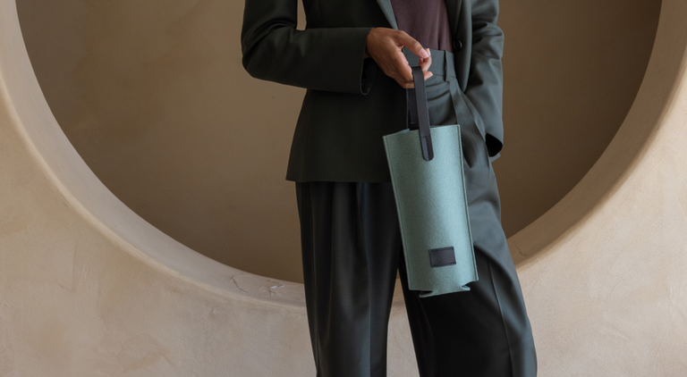 Person in denim outfit holding a brown felt wine carrier with a leather handle. They lean against a textured rock beside a wall with greenery.