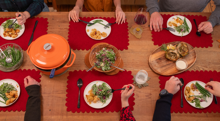 A table with four place settings features various pastries, a bowl of yogurt with fruit, glasses of water, and a persons arm reaching for food. Plates are on brown placemats.