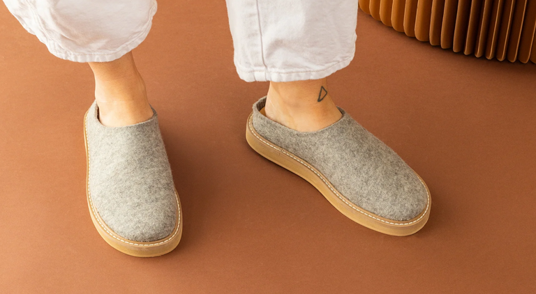 A person wearing cream socks and gray felt slip-on clogs with tan soles, paired with blue jeans, sitting on a light-colored surface.