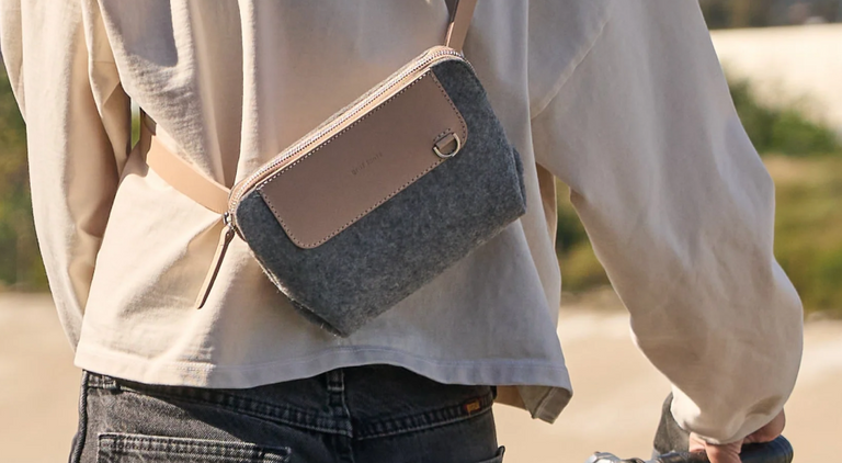 A person walking with a large, stylish black tote bag with geometric design, wearing black pants and wooden-heeled sandals. Sunlight casts shadows on a textured wall in the background.