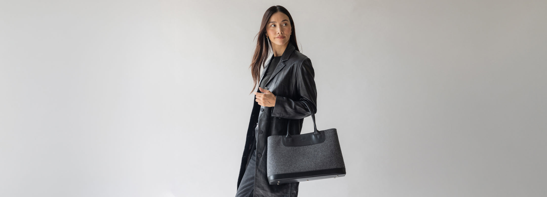 A person carrying a large, structured black tote bag with black handles. The person is wearing black pants and heeled sandals, walking against a sunlit concrete wall.